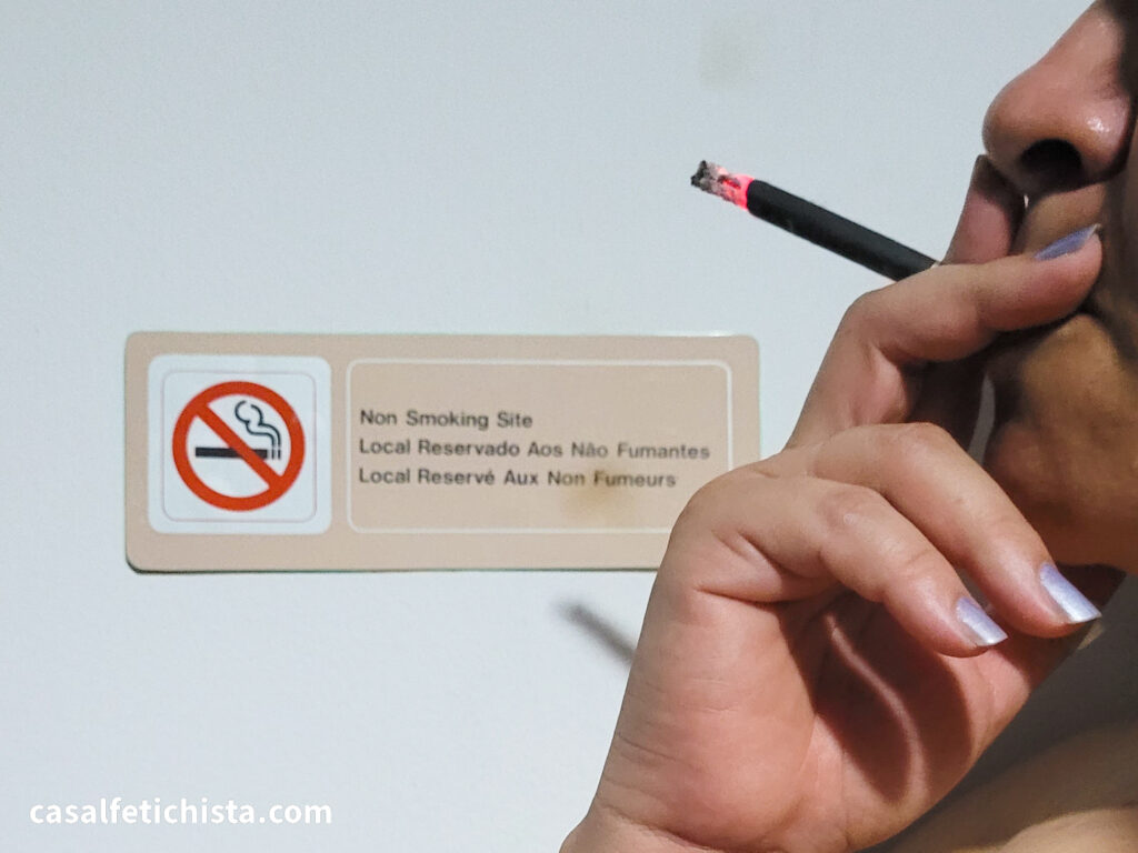 Mulher segurando cigarro aceso em frente a aviso de proibição de fumo